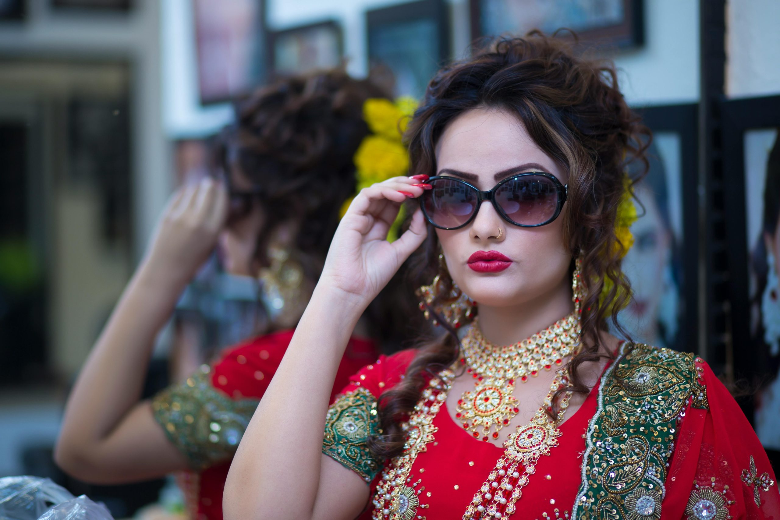Beautiful South Asian bride in traditional red attire, adorned with jewelry and sunglasses, posing indoors.