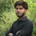 A young South Asian man poses confidently outdoors among greenery.