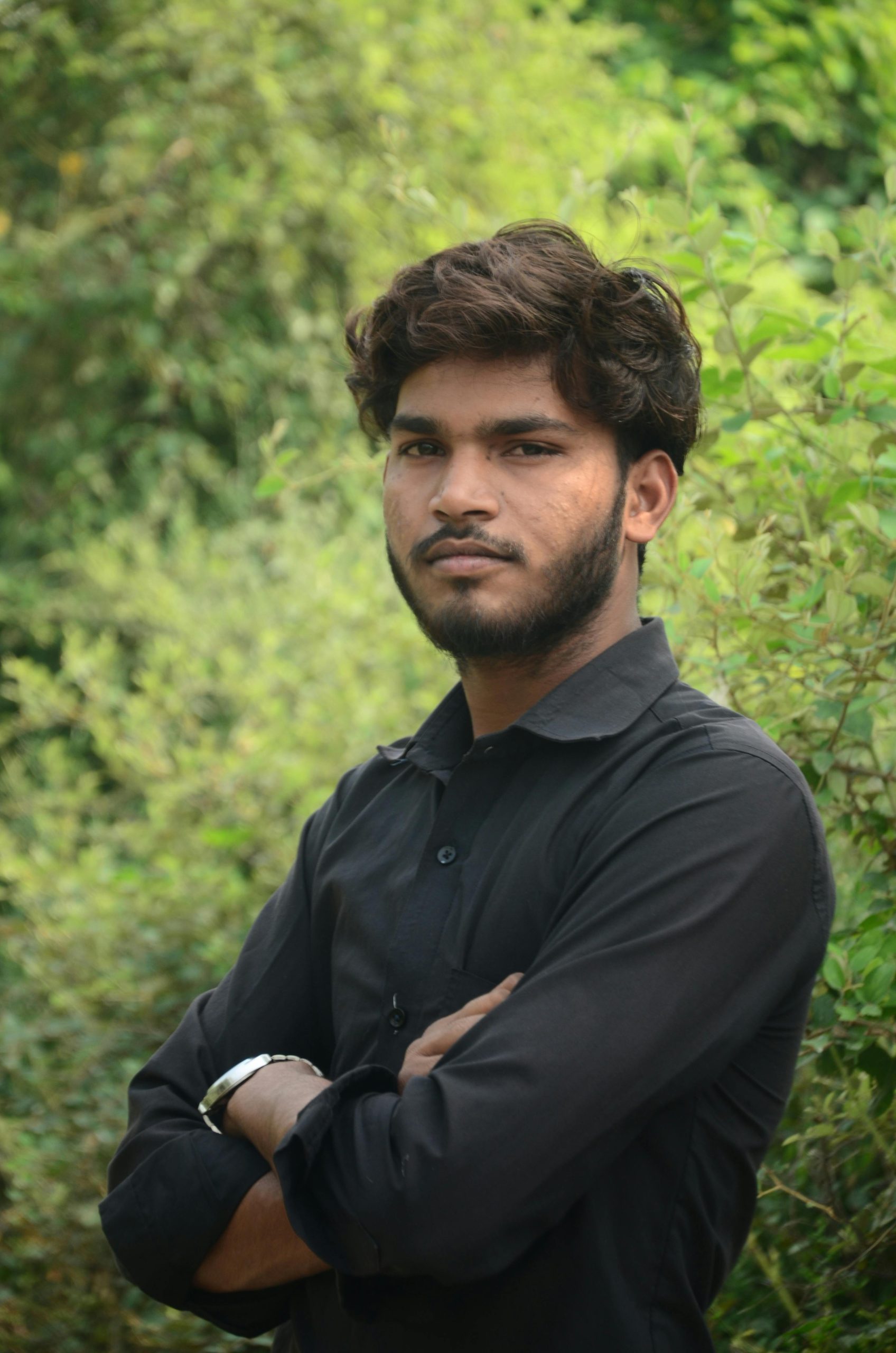 A young South Asian man poses confidently outdoors among greenery.